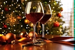 Kerstkaart: Twee glazen rode wijn staan op tafel met ernaast drie kerstballen en op de achtergrond een grote kerstboom